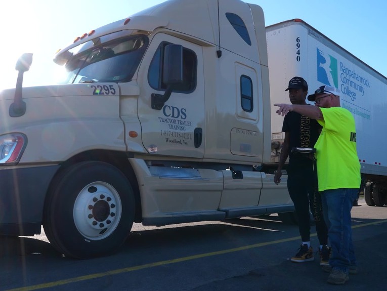 A CDS Tractor Trailer Training instructor in a neon yellow shirt points while coaching a student beside a tan semi truck. The truck's trailer carries Rappahannock Community College and Germanna Community College logos and a "Student Driver" label.